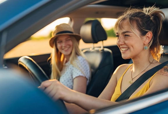 Two people seated in a blue vehicle during golden hour, with the driver holding the steering wheel and both occupants visible through the open side windows, illustrating vehicle ownership and automotive financing.