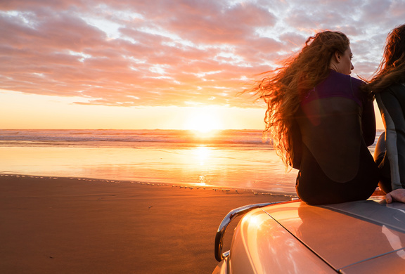 Two people sitting on a car at the beach watching the sunset.
