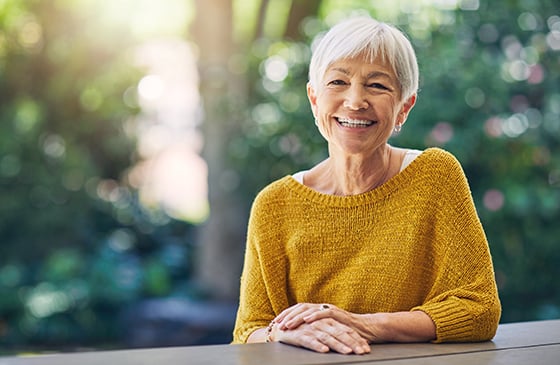 Older woman sitting outside at at bench smiling. 