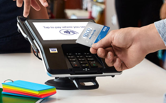A hand using a SELCO Visa card at a payment terminal in a store. 