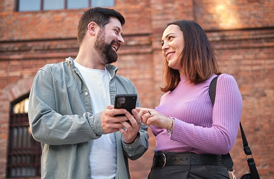 Two people using a smartphone together outside a brick building, representing the benefits of linking Link Digital Checking and Cultivate High-Yield Savings.
