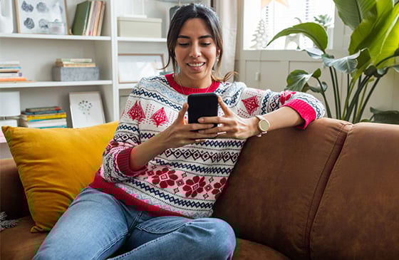 Woman sitting on the couch in a holiday sweater looking at her phone. 
