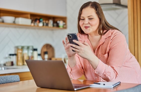 Lady smiling and using her phone with her laptop open in front of her.